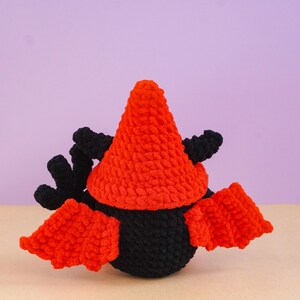 May include: A crocheted plush toy featuring a black rounded body, a bright orange pointed hat, and two orange wings extending from the sides. The toy also has black textured appendages protruding from the top, all crafted from soft yarn.