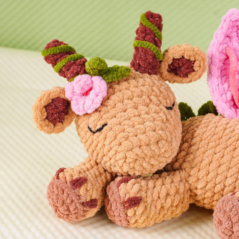 May include: A handmade, tan-colored crochet deer plushie with closed eyes, adorned with pink and green floral accents. The deer has brown and green antlers, and brown accents on its ears and hooves. The plushie is made of soft, textured yarn.
