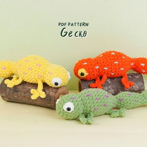 May include: Three crocheted gecko toys in yellow, orange, and green, each with contrasting spots and black and white eyes. The toys are posed on wooden logs. The text "PDF Pattern Gecko" is at the top.