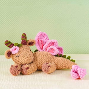 May include: A handmade, plush dragon toy in shades of brown, pink, and green. The dragon features pink wings, a flower on its head, and a curled tail. The toy is made of soft, textured yarn, perfect for a child's comfort.