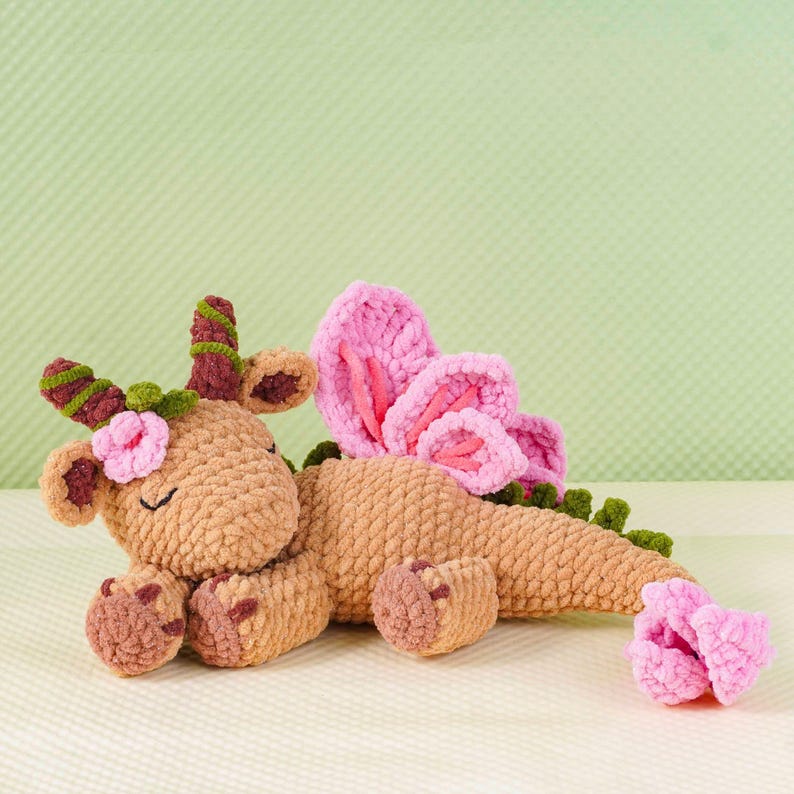 May include: A handmade, plush dragon toy in shades of brown, pink, and green. The dragon features pink wings, a flower on its head, and a curled tail. The toy is made of soft, textured yarn, perfect for a child's comfort.