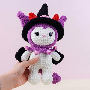 May include: A handmade crochet doll in the shape of a witch's familiar. The doll is primarily white with purple accents, including a hat, ears, and collar. The hat is black with red and pink details. The doll has black eyes and a pink nose.