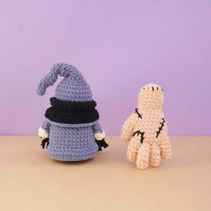 May include: A crochet gnome with a blue pointed hat, a black collar, and blue body, positioned next to a beige crochet mummy figure featuring black stitched details across its surface. Both items are crafted from thick, textured yarn.