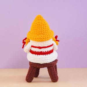 May include: A crocheted gnome figure featuring a tall, textured yellow hat, a white body with horizontal red stripes, and two red braided accents on the sides. The figure is seated on a small, dark brown crocheted stool with four legs.