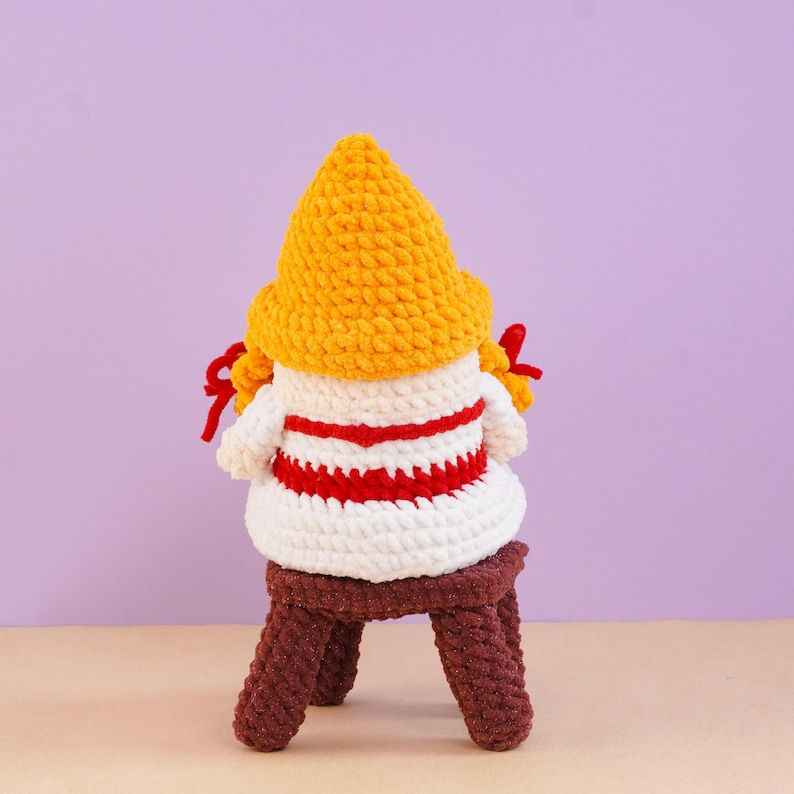 May include: A crocheted gnome figure featuring a tall, textured yellow hat, a white body with horizontal red stripes, and two red braided accents on the sides. The figure is seated on a small, dark brown crocheted stool with four legs.