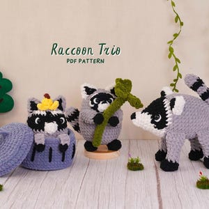 May include: Three crocheted raccoon figures with grey, white, and black yarn. One raccoon sits in a blue lidded basket, one holds a green leafy branch, and one stands on four legs. The text Raccoon Trio PDF PATTERN is displayed above them.