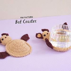 May include: Two crocheted bat coasters in tan and dark brown yarn. One coaster lies flat, while the other supports a clear glass mug. The text "PDF PATTERN Bat Coaster" is printed above the items. The coasters feature round bodies, small ears, and dark brown wings.