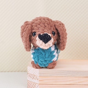 Puede incluir: Un dachshund de crochet marr&oacute;n que lleva un su&eacute;ter a rayas azul y blanco. El dachshund est&aacute; sentado sobre una superficie de madera.