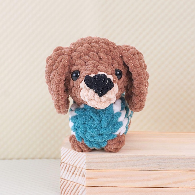 Puede incluir: Un dachshund de crochet marr&oacute;n que lleva un su&eacute;ter a rayas azul y blanco. El dachshund est&aacute; sentado sobre una superficie de madera.