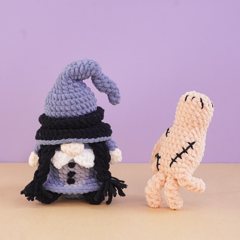 May include: A crochet gnome with a tall blue hat, black brim, white beard, and black braids, standing next to a beige crochet mummy figure featuring black stitched details and a textured, yarn-based construction throughout both items.