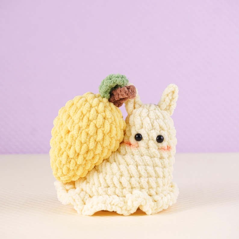 May include: A handmade crochet snail toy with a yellow shell and a cream-colored body. The snail has black eyes, rosy cheeks, and a small green and brown detail on its head. The toy is made of soft, textured yarn.
