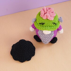 May include: A crocheted plush figure featuring a green head with a pink top, purple body, and gray beard, accompanied by a separate black crocheted hat. The figure has black stitched eyes and small gray side bolts, crafted from soft yarn.