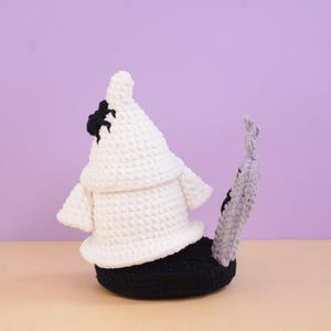 May include: A crocheted plush figure featuring a white ghost-like shape with a black spider on its side, positioned atop a black base. A tall, thin, light gray textured stalk with a small black detail extends upward from the black base.
