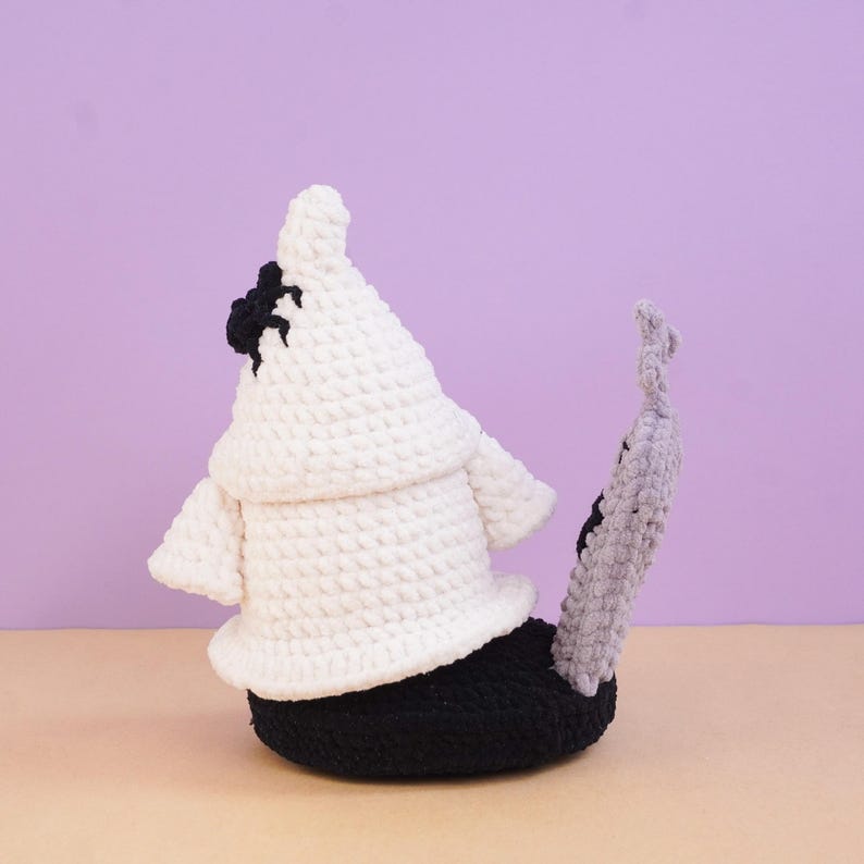 May include: A crocheted plush figure featuring a white ghost-like shape with a black spider on its side, positioned atop a black base. A tall, thin, light gray textured stalk with a small black detail extends upward from the black base.