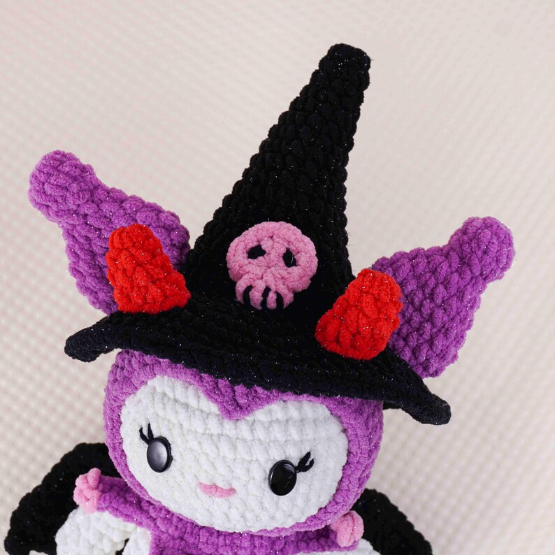May include: Crocheted plush toy in the shape of a bat-like creature, wearing a black witch hat with a pink skull. The toy has purple wings, red accents, and a white face with black eyes. A handmade Halloween decoration.