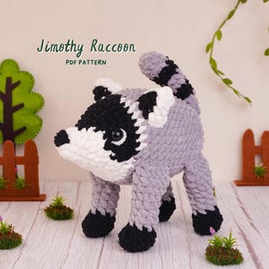 May include: A crocheted raccoon toy with grey, black, and white yarn, featuring a striped tail, black eye patches, and a white muzzle. The text Jimothy Raccoon PDF PATTERN is visible above the toy, which stands on four black-tipped legs.
