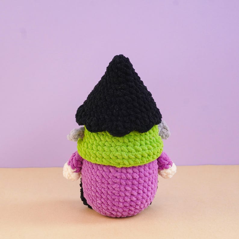 May include: A crochet plush figure featuring a tall, pointed black hat, a bright lime green textured midsection, and a purple base. The figure has small cream-colored hands extending from the sides and gray tufts of yarn near the hat brim.