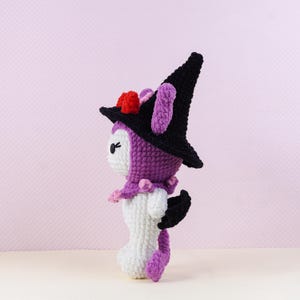 May include: A handmade crochet doll in a witch costume. The doll is primarily white, purple, and black, with a pointed black hat adorned with red and pink accents. It has a small, bat-like wing and a long, purple tail.