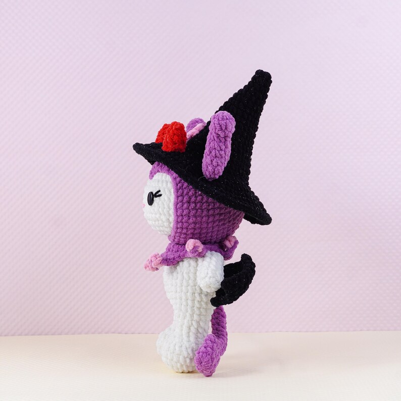 May include: A handmade crochet doll in a witch costume. The doll is primarily white, purple, and black, with a pointed black hat adorned with red and pink accents. It has a small, bat-like wing and a long, purple tail.