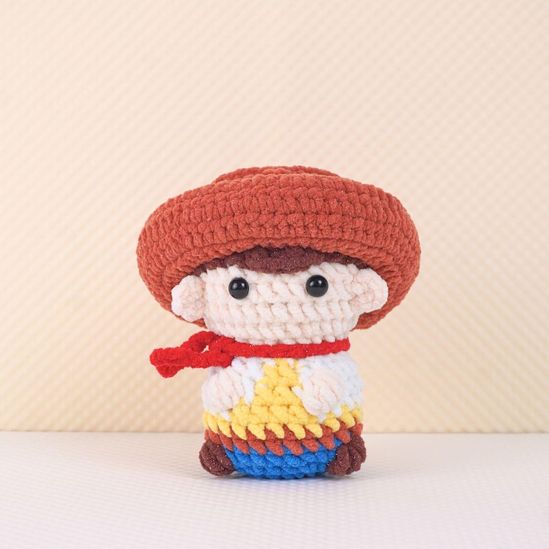 May include: A crocheted toy cowboy doll with a red bandana, brown hat, and colorful vest. The doll has a white face with black eyes and a brown mouth.
