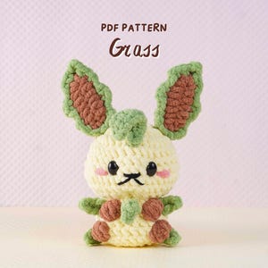 Könnte beinhalten: Ein gehäkeltes Spielzeug-Kaninchen in Creme, Braun und Grün. Das Kaninchen hat große Ohren, rosige Wangen und einen kleinen schwarzen Mund. Der Text "PDF PATTERN Grass" steht oben.