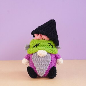 May include: A crocheted plush doll featuring a black pointed hat, a green textured head with black stitched details, a white nose, a grey beard, a purple body, and black feet. The soft yarn construction creates a rounded, stuffed appearance.