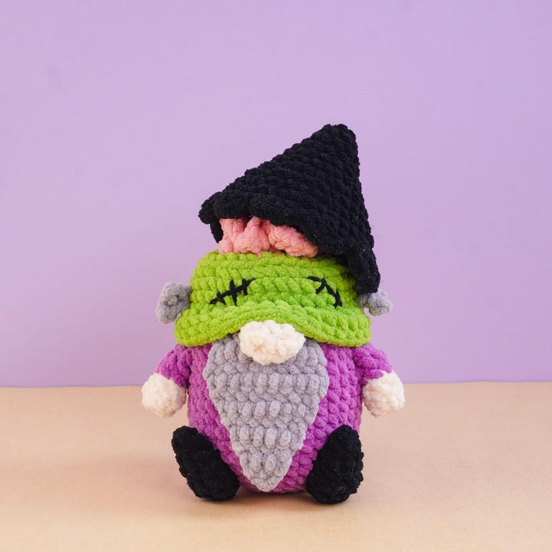 May include: A crocheted plush doll featuring a black pointed hat, a green textured head with black stitched details, a white nose, a grey beard, a purple body, and black feet. The soft yarn construction creates a rounded, stuffed appearance.