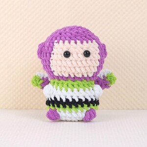 May include: A purple and green crocheted toy resembling a space ranger character. The toy has black eyes and a white face.