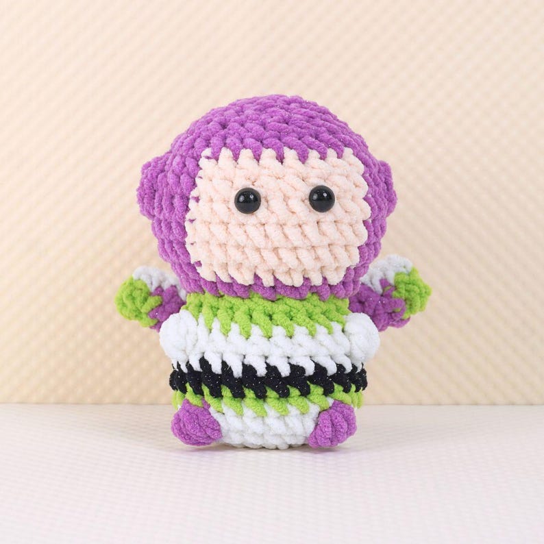 May include: A purple and green crocheted toy resembling a space ranger character. The toy has black eyes and a white face.
