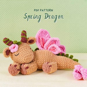 Puede incluir: Un juguete de dragón de ganchillo en tonos beige, rosa y verde. El dragón tiene detalles de flores rosas, grandes alas rosas y el texto "PDF PATTERN Spring Dragon" en un estilo manuscrito.