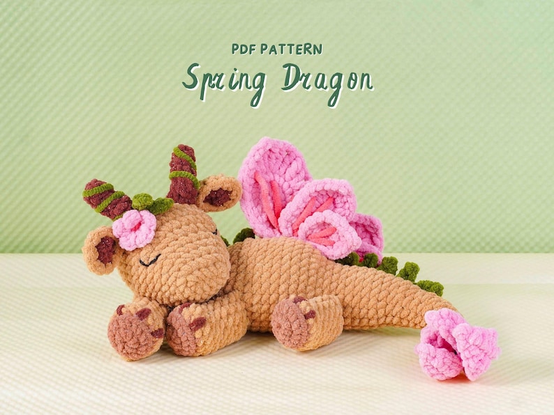 May include: A crocheted dragon toy in shades of tan, pink, and green. The dragon has pink flower accents, large pink wings, and the text "PDF PATTERN Spring Dragon" in a handwritten style.