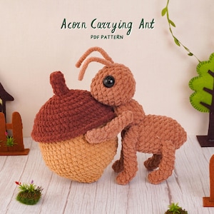 May include: A crocheted light brown ant with black eyes holding a large crocheted acorn with a dark brown cap and tan base. The text Acorn Carrying Ant PDF PATTERN is visible at the top. The ant has two antennae and six legs.