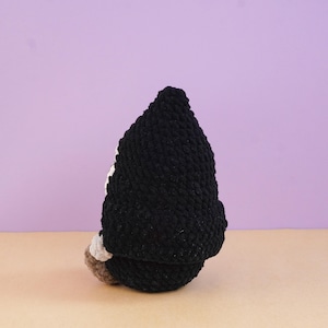May include: A handmade crochet gnome figurine featuring a tall, pointed black hat made of textured yarn. The gnome has a small visible portion of a white beard and a tan-colored base, showcasing a soft, plush, and rounded construction.
