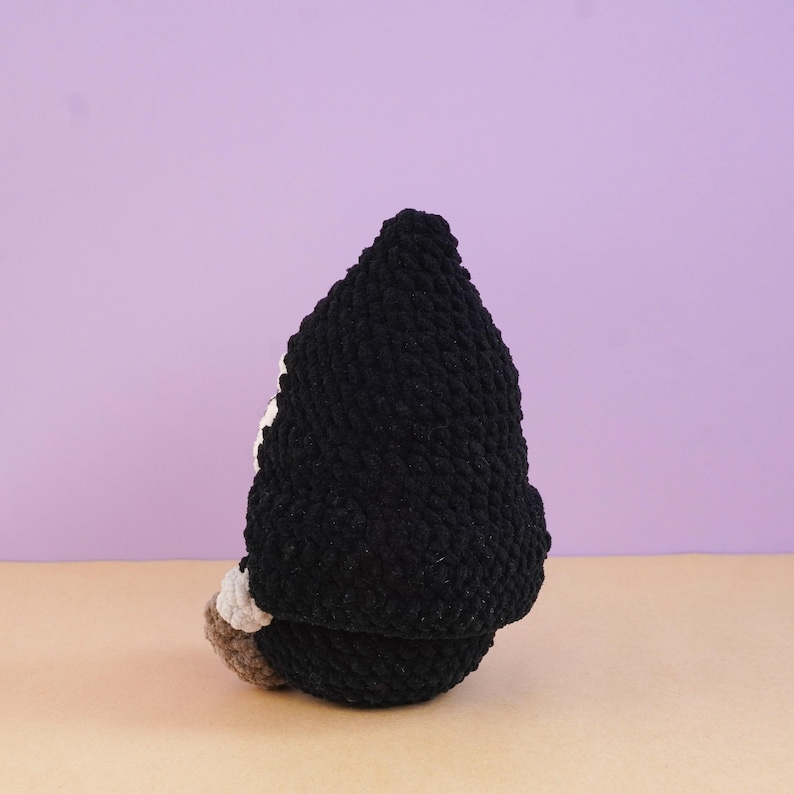 May include: A handmade crochet gnome figurine featuring a tall, pointed black hat made of textured yarn. The gnome has a small visible portion of a white beard and a tan-colored base, showcasing a soft, plush, and rounded construction.