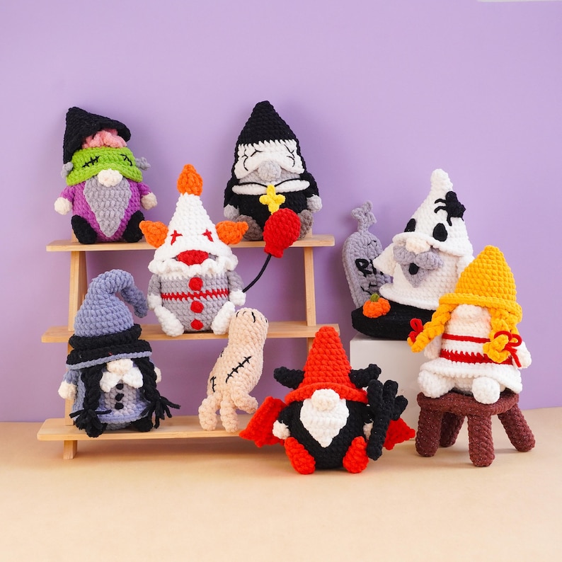 May include: A collection of eight crocheted Halloween-themed gnomes and figures, including characters resembling a clown with a red balloon, a vampire, a ghost with a spider, a mummy, and a figure with braided yellow hair, all crafted in yarn.