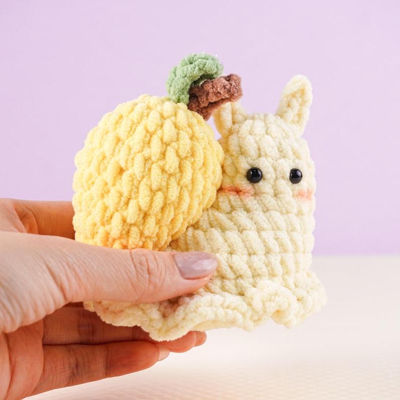 May include: A handmade crochet snail toy in soft yellow and cream colors. The snail has a textured shell, a cream-colored body with black eyes and rosy cheeks, and a small green and brown accent on top. The toy is held in a hand.