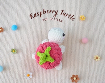 May include: A crocheted raspberry turtle amigurumi toy with a white shell and a pink raspberry body. The toy is on a white fuzzy background with small flowers.