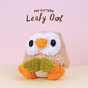 May include: A crocheted owl with a tan body, white face, orange beak, and black eyes. The owl is holding green and orange leaves. The text "PDF PATTERN Leafy Owl" is above the owl.