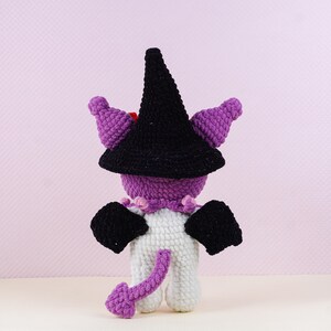 May include: A handmade crochet doll in a witch costume. The doll has a black witch hat, purple horns, and black wings. The body is white with a purple tail. The doll is wearing a purple collar with pink accents.
