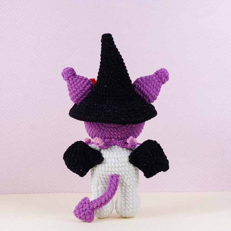 May include: A handmade crochet doll in a witch costume. The doll has a black witch hat, purple horns, and black wings. The body is white with a purple tail. The doll is wearing a purple collar with pink accents.