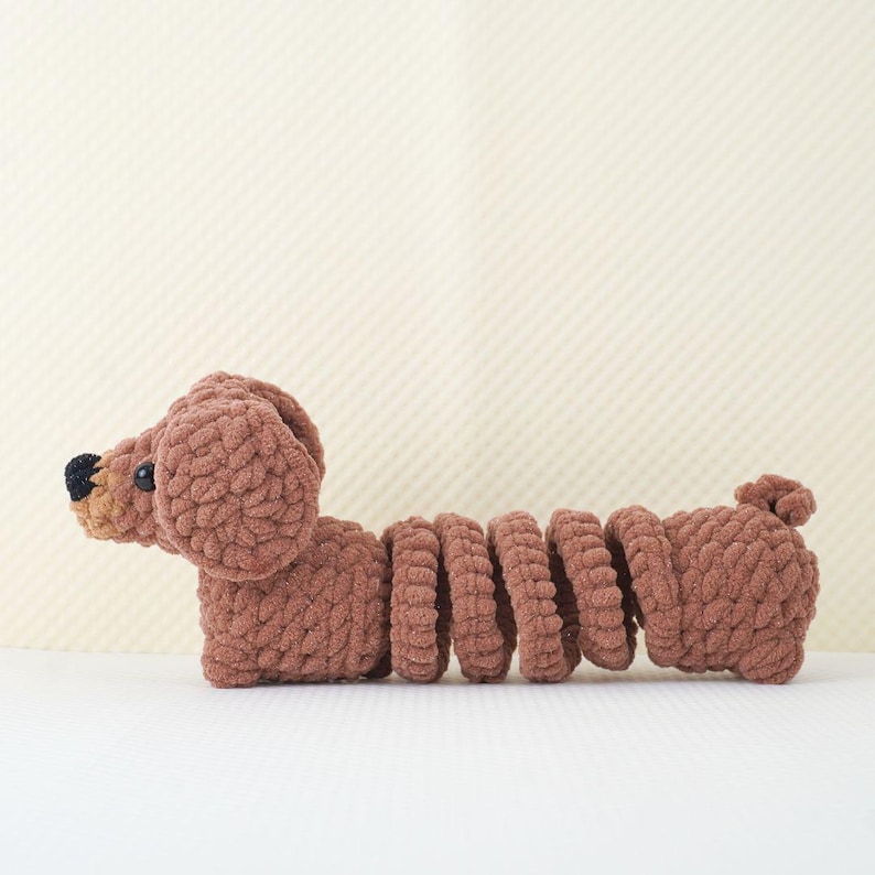 Puede incluir: Un juguete de peluche de dachshund de color marr&oacute;n tejido a crochet con ojos negros y cola enroscada. El juguete est&aacute; hecho de hilo suave y es perfecto para acurrucarse.