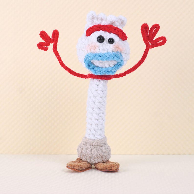 May include: A white crocheted toy with red arms, a blue mouth, and brown feet. The toy is shaped like a person and has a red headband.