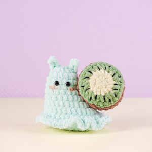 May include: A handmade crochet snail toy in light blue with a kiwi shell. The snail has black eyes and blush-colored cheeks. The kiwi shell is green, brown, and cream-colored. The toy is on a light purple background.
