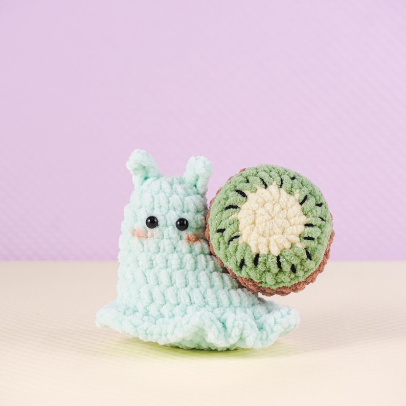 May include: A handmade crochet snail toy in light blue with a kiwi shell. The snail has black eyes and blush-colored cheeks. The kiwi shell is green, brown, and cream-colored. The toy is on a light purple background.