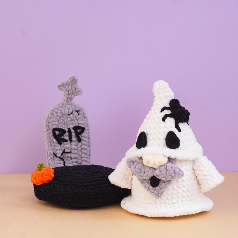 May include: A crocheted white ghost figure with a black spider on its head and a gray beard, positioned next to a gray crocheted tombstone with the letters RIP embroidered in black, sitting on a black base with a small orange pumpkin.
