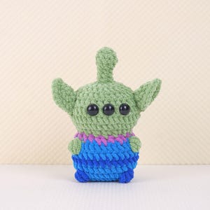May include: A green crocheted alien plush toy with three black eyes and a blue and purple body.