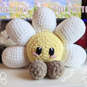 Puede incluir: Un juguete de peluche de margarita tejido a crochet con un centro amarillo y pétalos blancos. La flor tiene una cara linda con ojos negros y mejillas rosadas. El juguete tiene dos bolas marrones tejidas a crochet para las piernas.