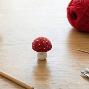 Könnte beinhalten: Ein handgehäkelter Pilz mit rotem Hut und weißen Punkten, auf einer Holzoberfläche. Bastelmaterialien, darunter rote Wolle, eine Häkelnadel und eine Schere, sind zu sehen. Ein charmantes, handgefertigtes Dekorationsobjekt.