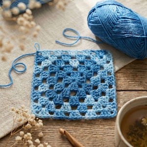 May include: A square, blue and white crocheted granny square sits on a wooden surface, with a ball of blue yarn and a crochet hook nearby. The square is made of varying shades of blue yarn, creating a textured pattern. A cup of tea and dried flowers are also in the image.