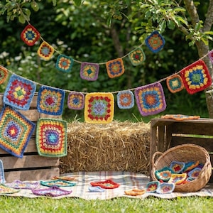 May include: A colorful crochet garland with square motifs in various colors hangs outdoors. The garland is strung above a wooden crate, a basket, and a hay bale. Additional crochet squares and coasters are arranged on a blanket on the grass.